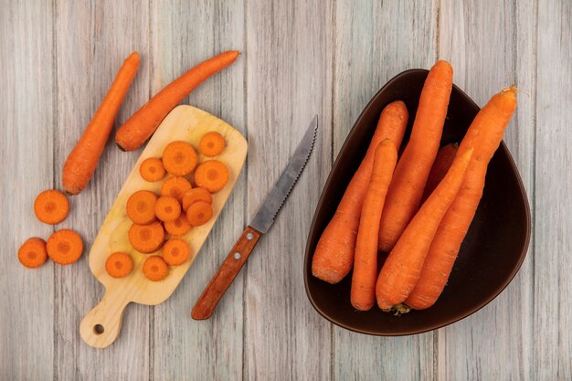 Vista superior de sabrosas zanahorias picadas en una tabla de cocina de madera con cuchillo con zanahorias en un recipiente sobre un fondo de madera gris