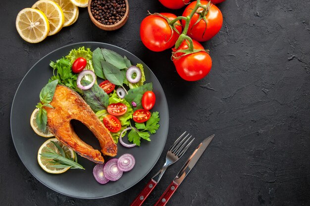 Vista superior sabroso pescado cocido con verduras frescas y cubiertos en una mesa oscura