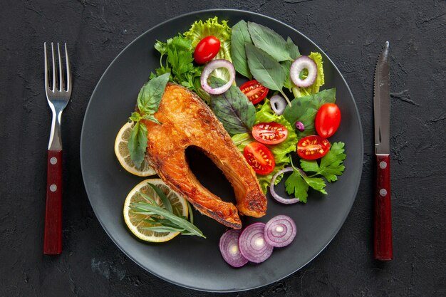 Vista superior sabroso pescado cocido con verduras frescas y cubiertos en una mesa oscura