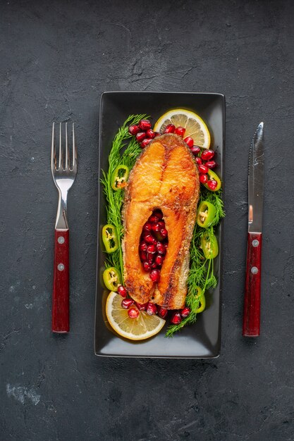 Vista superior sabroso pescado cocido con verduras y rodajas de limón dentro de la sartén en la mesa oscura