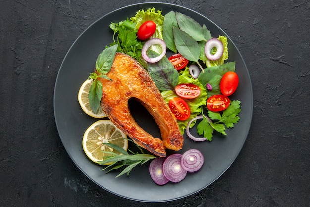 Vista superior sabroso pescado cocido con verduras y rodajas de limón en la mesa oscura