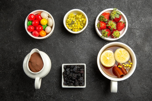 Vista superior del sabroso postre una taza de apetitoso té de hierbas junto a los tazones de crema de chocolate, coloridos caramelos, hierbas y fresas en la mesa oscura