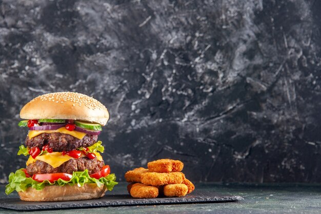 Vista superior del sabroso sándwich de carne con tomates verdes en bandeja de color oscuro y nuggets de pollo en el lado derecho sobre superficie negra