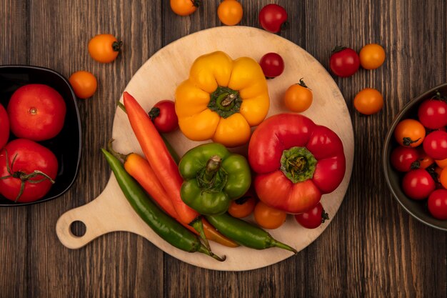 Vista superior de sabrosos pimientos en una tabla de cocina de madera con tomates frescos en un tazón en una pared de madera