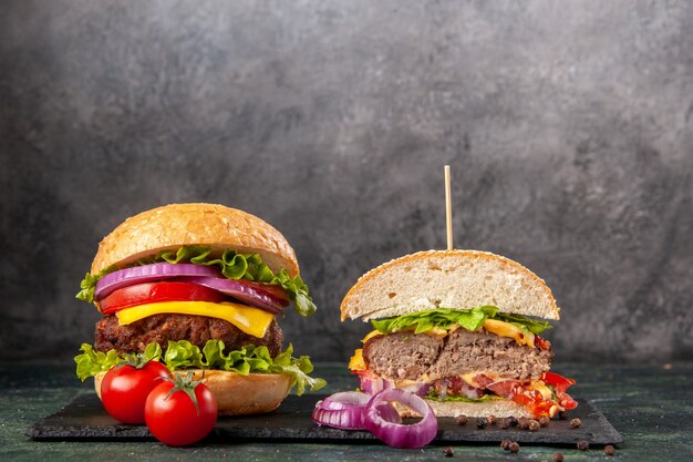 Vista superior de sándwiches y tomates sabrosos enteros cortados con cebollas de tallo en una bandeja negra sobre una superficie de color oscuro