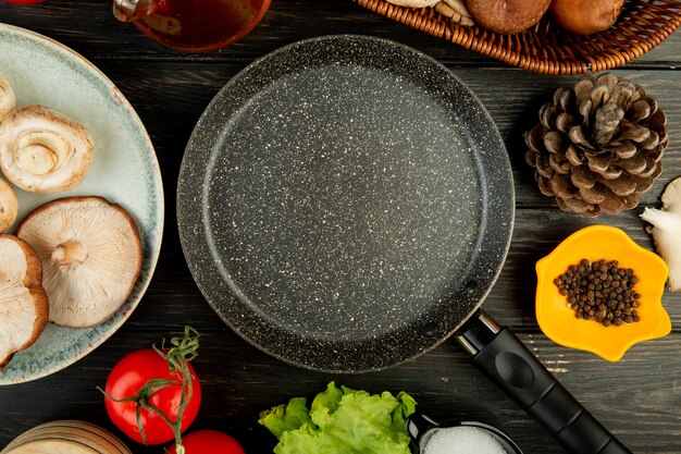 Vista superior de la sartén y champiñones frescos con conos de tomate granos de pimienta negra dispuestos alrededor de madera negra