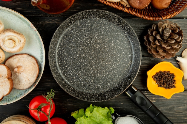 Vista superior de la sartén y champiñones frescos con conos de tomate granos de pimienta negra dispuestos alrededor de madera negra