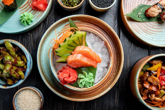 Vista superior de sashimi de salmón con pepinos en rodajas jengibre y salsa de wasabi en cubitos de hielo en un recipiente sobre la mesa de madera