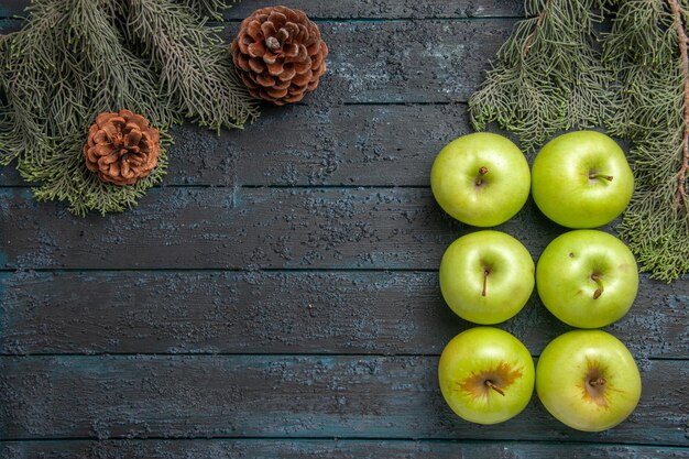Vista superior seis manzanas seis apetitosas manzanas verdes en el lado derecho de la mesa gris junto a las ramas de los árboles con conos