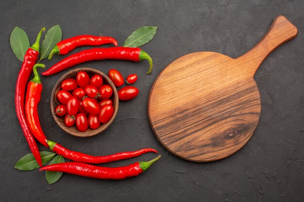 Vista superior del semicírculo de pimientos rojos y hojas de laurel y un tazón de tomates cherry y una tabla de cortar ovalada sobre la mesa negra