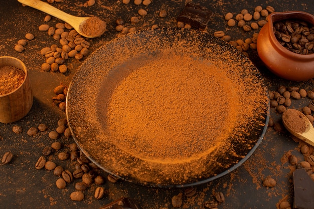 Una vista superior de las semillas de café marrón junto con una placa negra llena de café en polvo con barras de chocolate por toda la mesa marrón