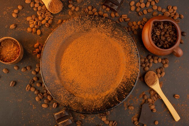 Una vista superior de las semillas de café marrón junto con una placa negra llena de café en polvo con barras de chocolate por todo el fondo marrón