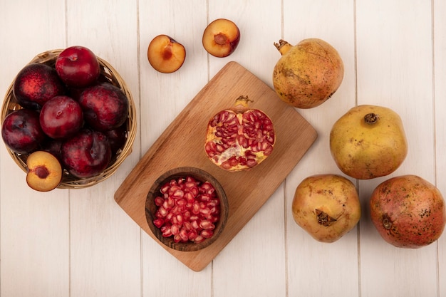 Foto gratuita vista superior de semillas de granada en un cuenco de madera sobre una tabla de cocina de madera con pluots en un balde con granadas aislado sobre un fondo blanco de madera