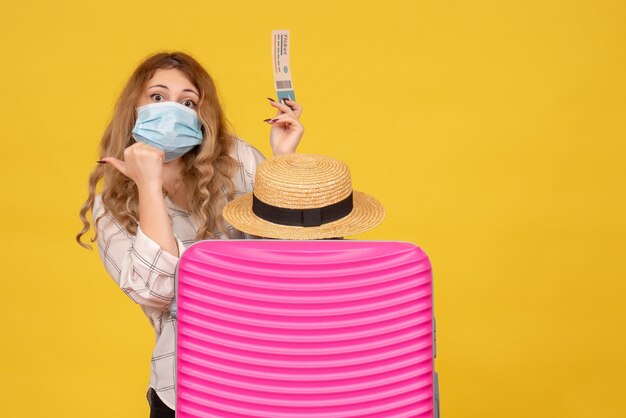 Vista superior de la señorita con máscara que muestra el boleto y de pie detrás de su bolso rosa apuntando hacia atrás