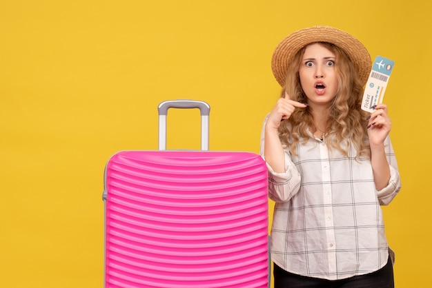 Vista superior de la señorita con sombrero sosteniendo boleto y de pie cerca de su bolso rosa apuntando a sí misma