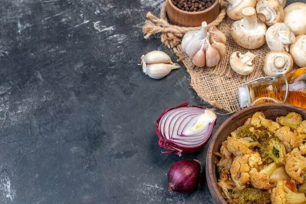 Vista superior de setas frescas, sabrosa coliflor en un tazón, botella de aceite, pimienta negra en un pequeño tazón de madera, ajo, cebolla roja en el lugar de copia de la mesa gris