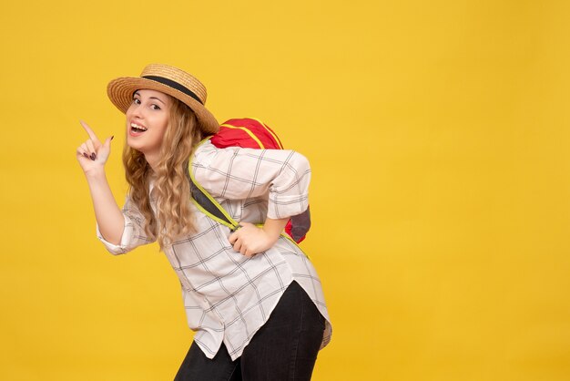 Vista superior de la sonriente chica viajera con sombrero y mochila roja apuntando hacia arriba en amarillo