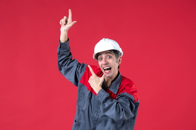 Vista superior del sonriente constructor joven feliz en uniforme con casco y apuntando hacia arriba en la pared roja aislada