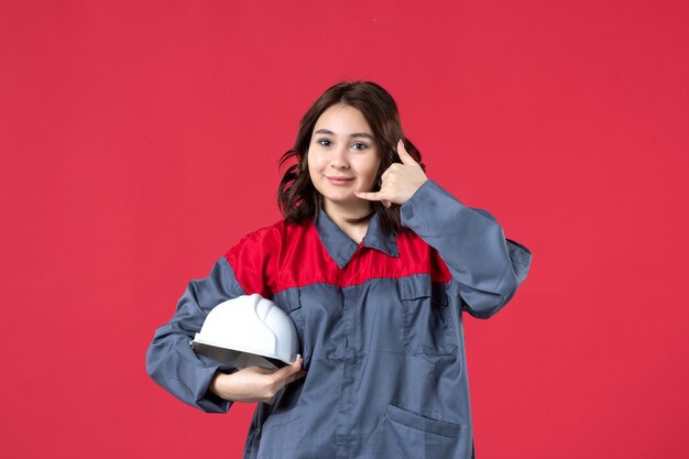 Vista superior de la sonriente constructora en uniforme y sosteniendo el casco haciendo gesto de llamarme sobre fondo rojo aislado