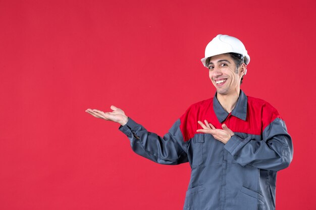 Vista superior del sonriente joven arquitecto confiado en uniforme con casco en pared roja aislada