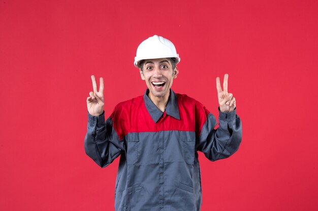 Vista superior del sonriente joven constructor en uniforme con casco haciendo gesto de victoria en la pared roja aislada