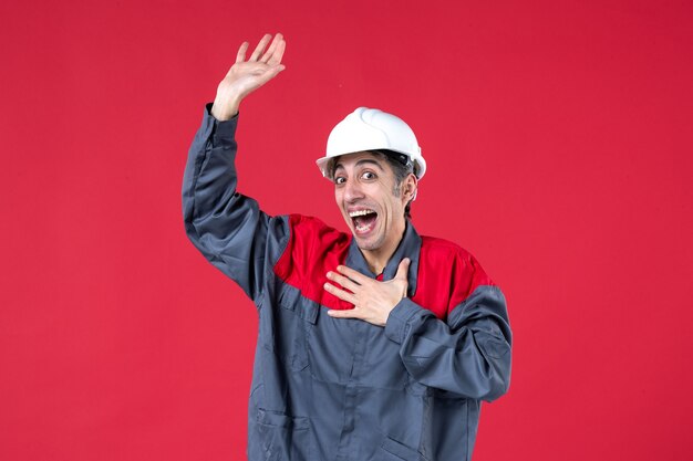 Vista superior del sonriente joven constructor en uniforme con casco sintiéndose confiado en la pared roja aislada