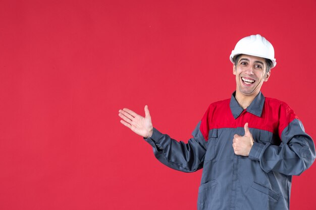 Vista superior del sonriente trabajador joven confiado en uniforme con casco y haciendo un gesto de ok en la pared roja aislada