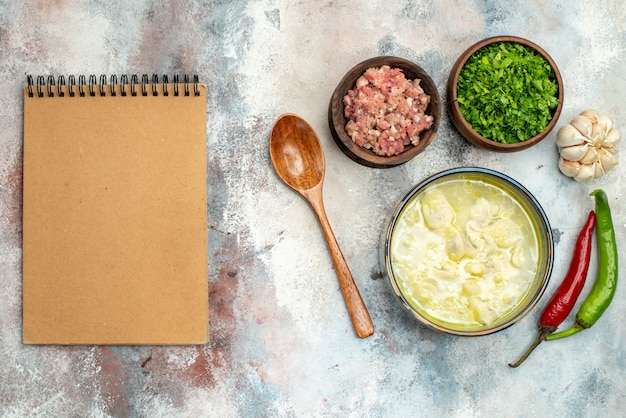 Vista superior sopa de albóndigas dushbara en un tazón de fuente de ajo pimientos picantes cuchara de madera con carne y verduras un cuaderno sobre una superficie desnuda