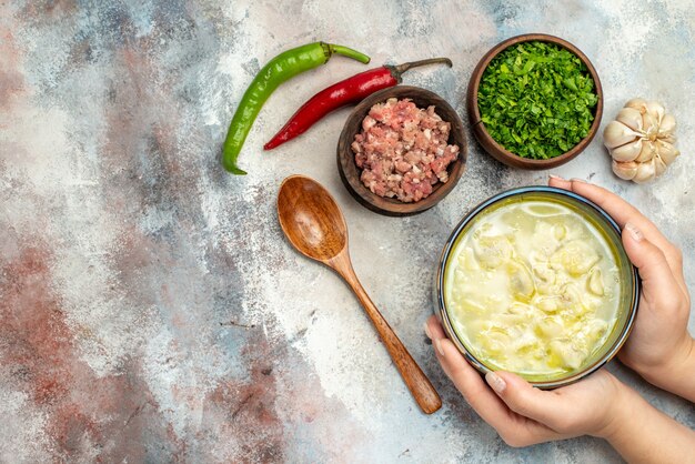 Vista superior de sopa de albóndigas de dushbara en un tazón en mano de mujer, pimientos picantes de ajo, cuencos de cuchara de madera con carne y verduras en una superficie desnuda con espacio de copia