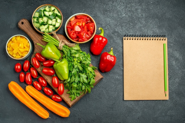 Vista superior del soporte para platos con verduras encima y cerca de él y el bloc de notas en el costado sobre fondo gris oscuro