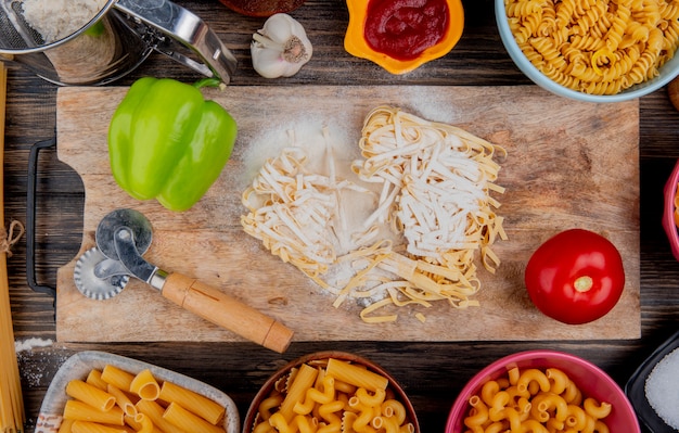 Vista superior de tagliatelle macarrones con harina, pimiento y tomate en la tabla de cortar con otros tipos de salsa de tomate salsa de ajo en la superficie de madera