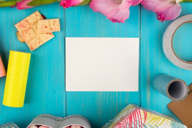 Foto gratuita vista superior de la tarjeta de felicitación de papel en blanco con flor de gladiolo de color rosa y chocolate blanco en la mesa de madera azul