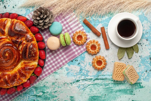 Vista superior de tarta de fresa con galletas macarons franceses y taza de té en superficie azul