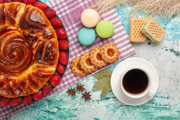 Vista superior de tarta de fresa con taza de té y macarons franceses en superficie azul