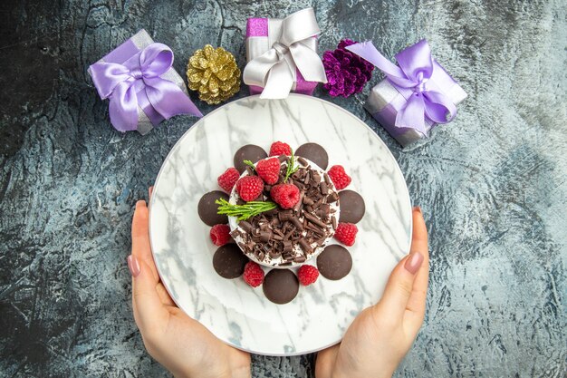 Vista superior de tarta de queso con chocolate con frambuesas en placa ovalada en manos de mujer regalos de navidad en espacio libre de superficie gris