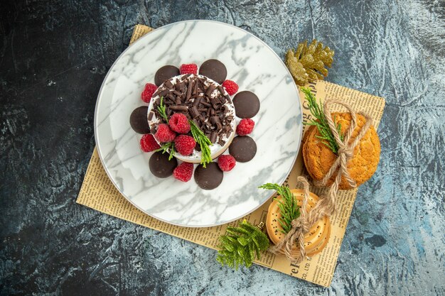 Vista superior de tarta de queso con chocolate y frambuesas en plato ovalado blanco galletas atadas en adornos navideños de periódico en superficie gris