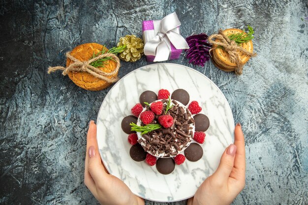 Vista superior de tarta de queso con chocolate en placa ovalada en manos de mujer galletas regalos de navidad en superficie gris espacio libre