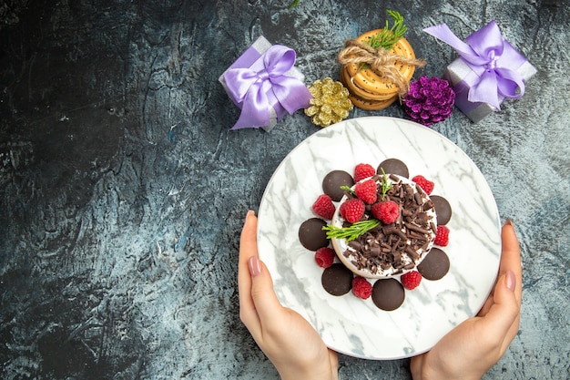 Vista superior de tarta de queso con chocolate en placa ovalada en manos de mujer regalos de navidad en espacio libre de superficie gris