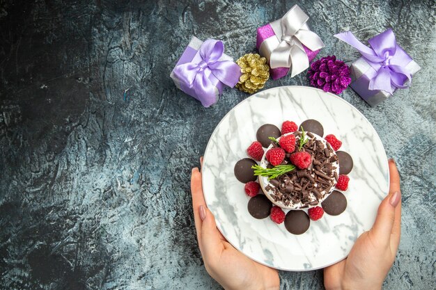 Vista superior de tarta de queso con chocolate en placa ovalada en manos de mujer regalos de navidad en lugar libre de superficie gris