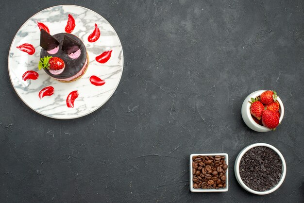 Vista superior de tarta de queso con fresas en un plato ovalado blanco en la parte superior izquierda y tazones con semillas de café de chocolate y fresas en la parte inferior derecha del fondo oscuro