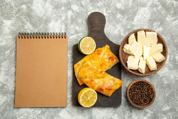 Vista superior de tartas, cuaderno de crema de queso junto a los tazones de tartas apetitosas de queso de pimienta negra y limón en la tabla de cortar en el lado derecho de la mesa