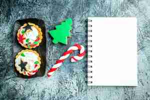 Foto gratuita vista superior de tartas de navidad en placa negra, dulces de navidad y bloc de notas de galletas de árbol de navidad en mesa gris