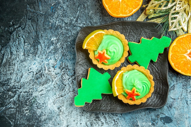 Vista superior tartas pequeñas con crema pastelera verde galletas de árbol de navidad en placa negra adorno de navidad naranjas cortadas en el espacio libre de la mesa gris