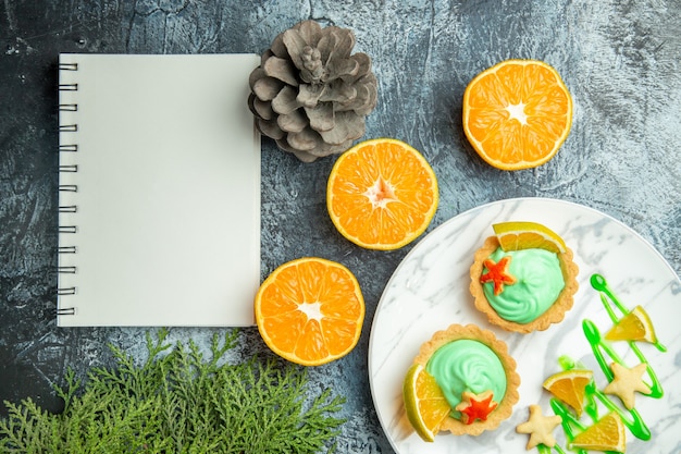 Vista superior de tartas pequeñas con crema pastelera verde y una rodaja de limón en un cuaderno de naranjas cortadas en placa en una mesa oscura