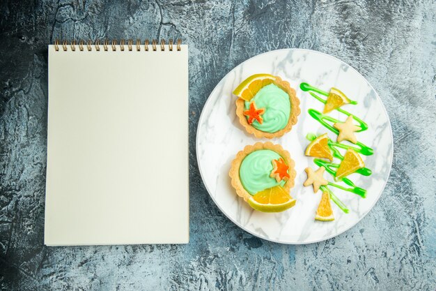 Vista superior de tartas pequeñas con crema pastelera verde y rodaja de limón en el cuaderno de placa en la mesa oscura