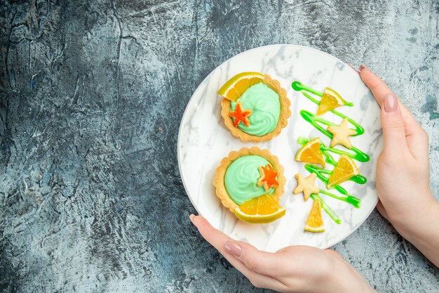 Vista superior de tartas pequeñas con crema pastelera verde y rodaja de limón en un plato en manos de mujer en la mesa oscura con lugar libre