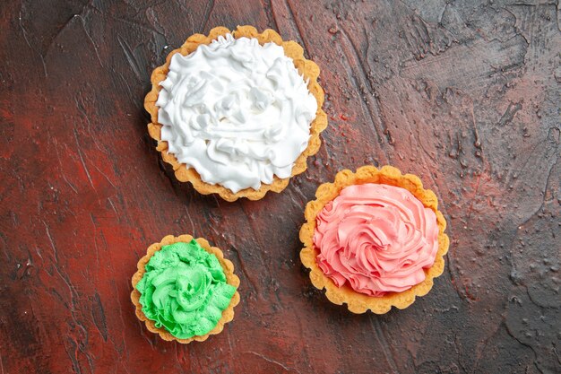 Vista superior de tartas pequeñas de diferentes tamaños con crema pastelera verde, rosa y blanca sobre una superficie de color rojo oscuro
