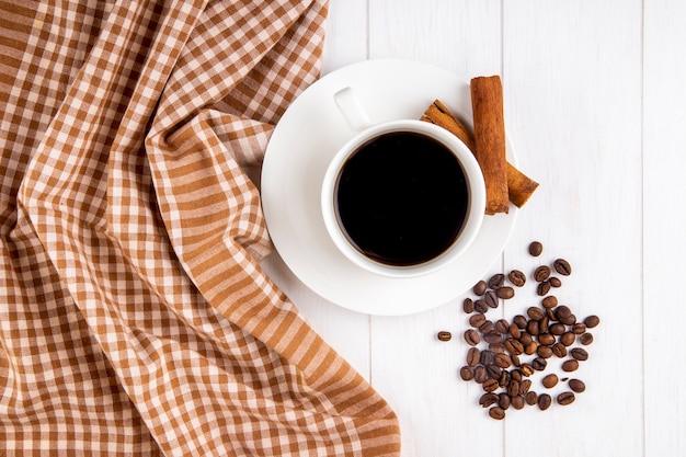 Vista superior de una taza de café con canela y granos de café esparcidos sobre fondo blanco de madera