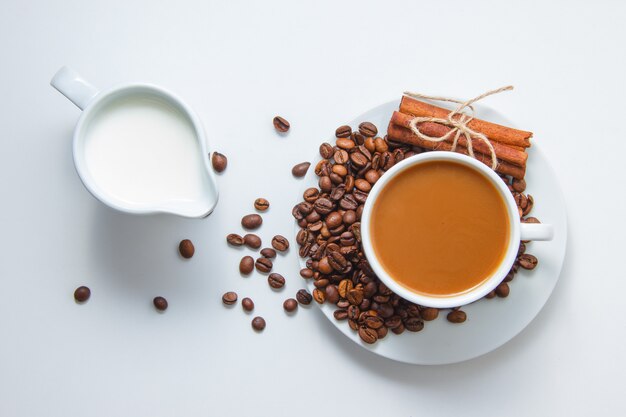 Vista superior de una taza de café con granos de café y canela seca en el plato y con leche, sobre una superficie blanca