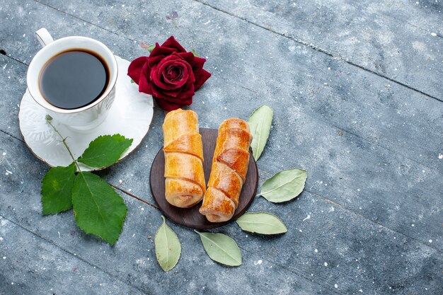 Vista superior de la taza de café junto con brazaletes deliciosos dulces y rosa roja en madera gris, azúcar de pastel de pastelería dulce hornear